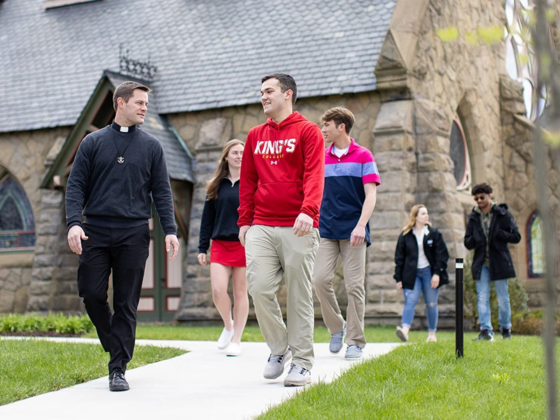 a priest walks with several students outside the Chapel