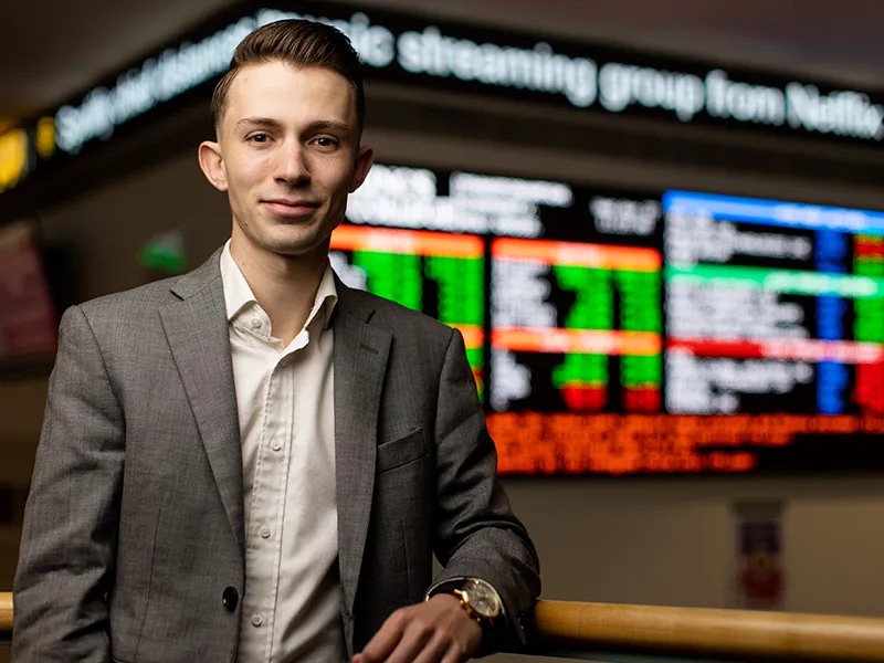 student in a suit poses in front of a stock ticker